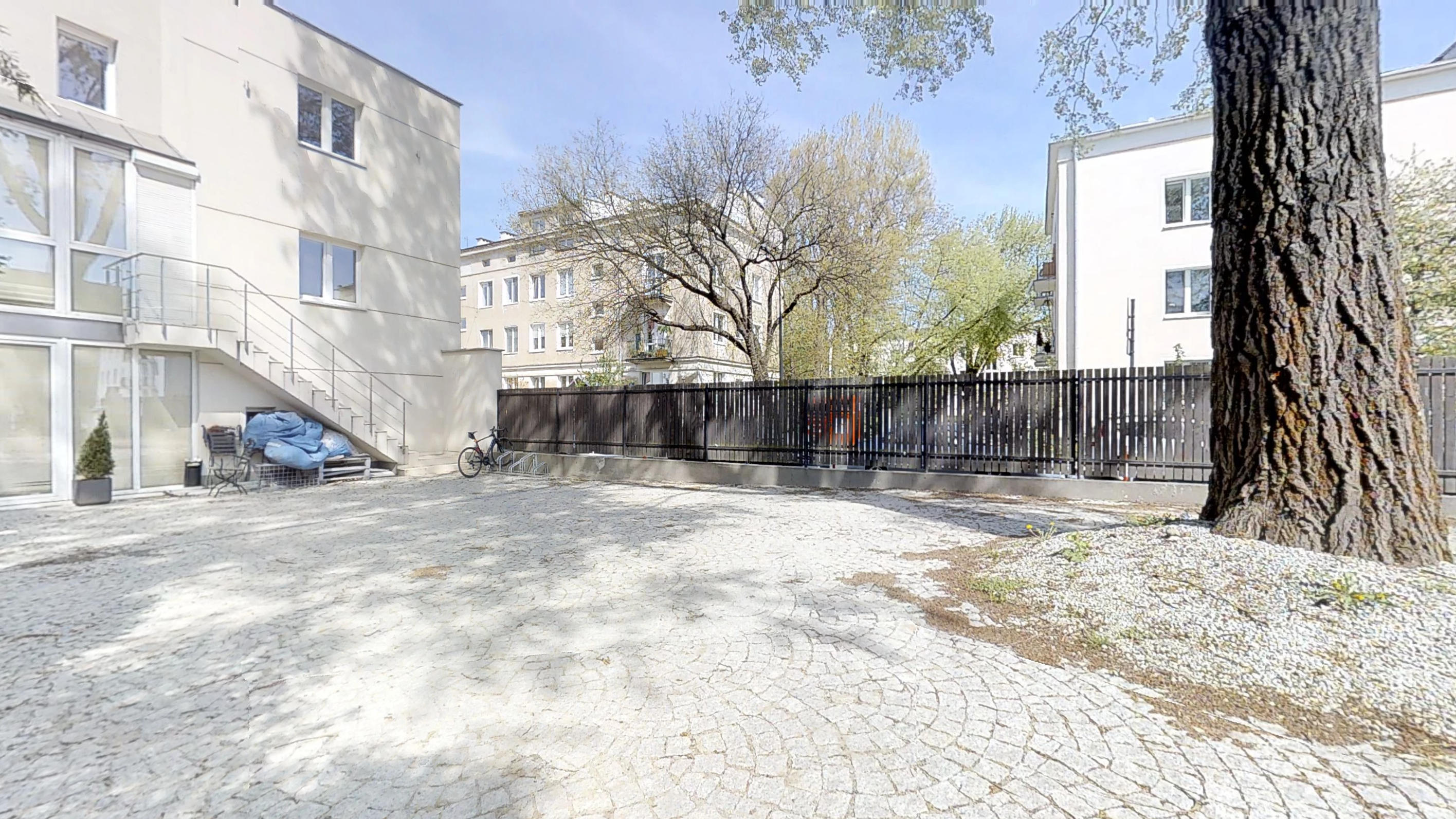 Utwardzony dziedziniec za budynkiem, duże drzewo po prawej i zewnętrzne schody przy fasadzie
